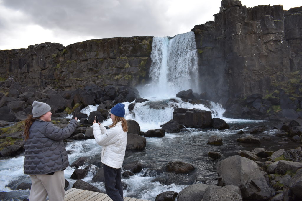 two women standing in front of a waterfall and taking pictures with their phones