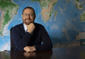 Man smiling with his hand on his chin sitting in front of a map of the world