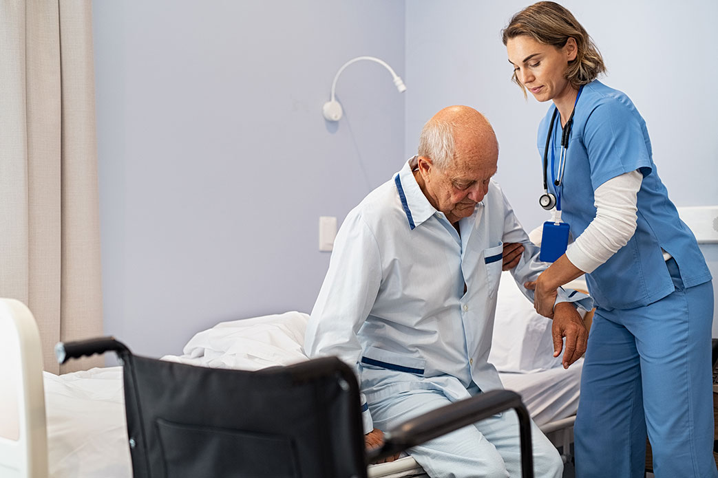 Nurse dressed in blue outfit helping an older gentleman into a wheelchair.