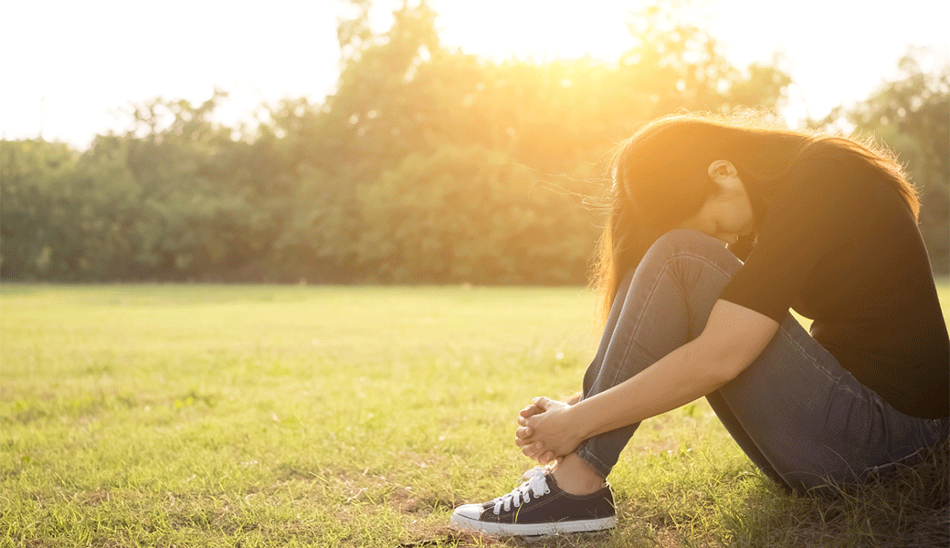Young female with her head tucked between her knees with feelings of depression sitting in an open field.