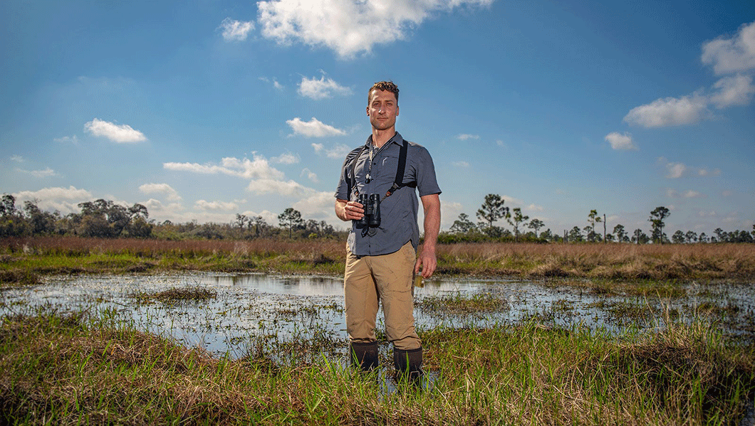 Man standing in a marshy field holding binoculars.