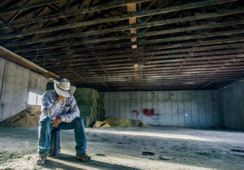 Farmer wearing a cowboy hat is sitting in a barn and leaning over to holding his hand over his face as a sign of depression.