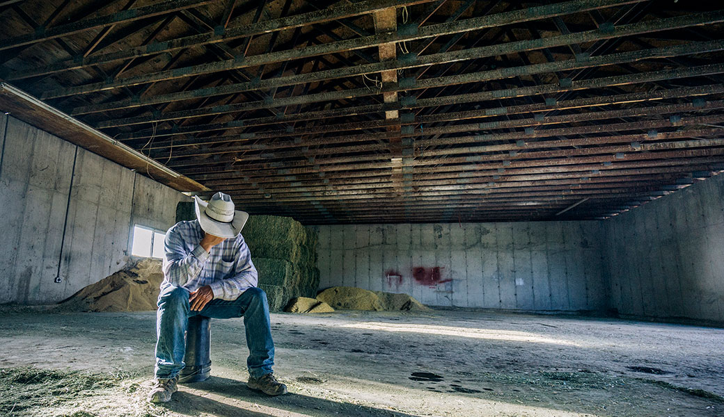 Farmer wearing a cowboy hat is sitting in a barn and leaning over to holding his hand over his face as a sign of depression.