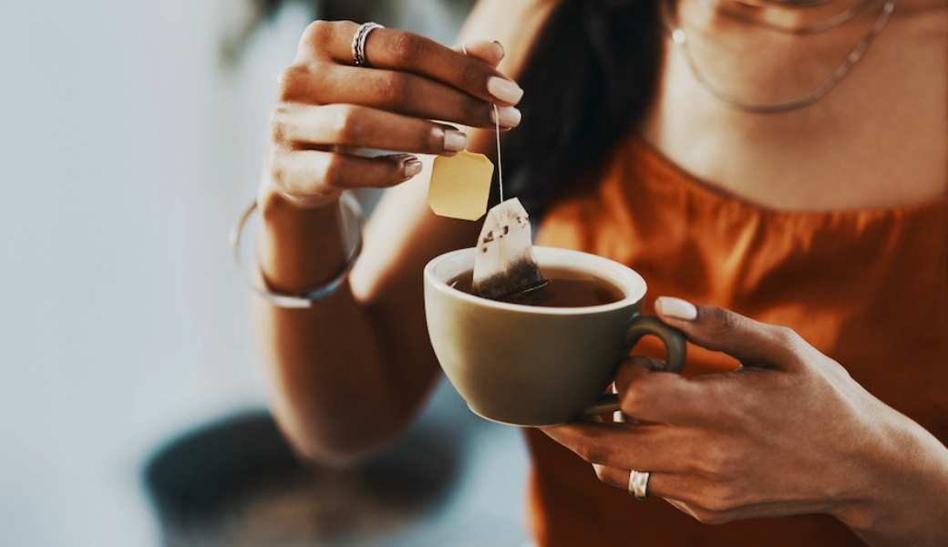 A woman holding a cup of tea in one hand, and a teabag submerged in the cup in her other hand.