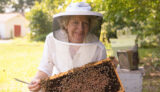 Woman smiling in a beekeeper suit holding up a honeybee hive from a bee box.