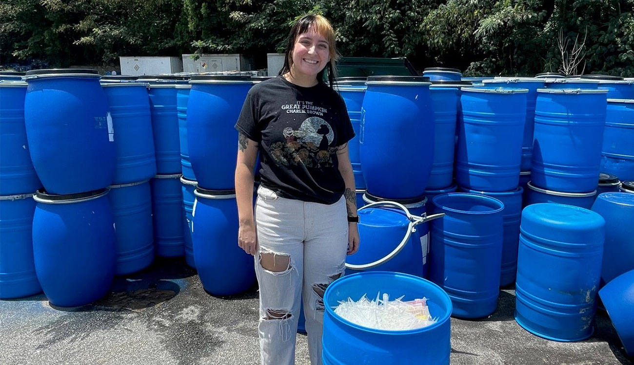 A student worker stands in front of blue plastic barrels. A blue plastic barrel filled with pipette tips sits to the right of the student worker.