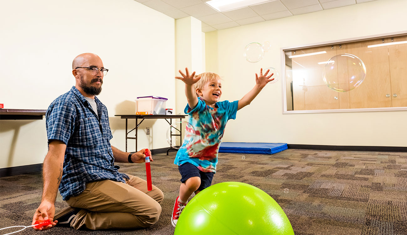 A man oversees a smiling toddler playing in a classroom