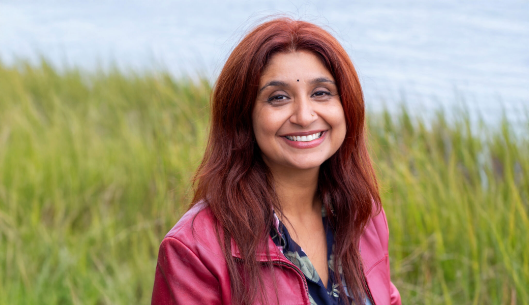 Image of Mona Behl wearing a pink jacket and blue blouse standing in a field of grass next to a body of water.