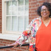 Barbara McCaskill stands in front of a brick wall wearing an orange top, patterned jacket, and glasses.