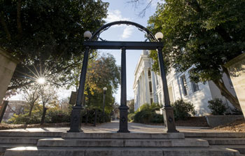 A black iron archway stands at the top of stone steps, framed by trees and a white building in the background under a partly cloudy sky.