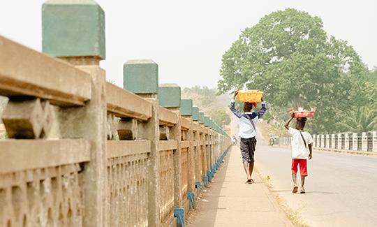Children in Cambia crossing over a bridge and holding items on top of their heads as they walk.